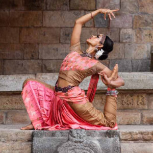 Woman in colorful pink Dance attire sits with her back arched and arms flared out on a stone surface
